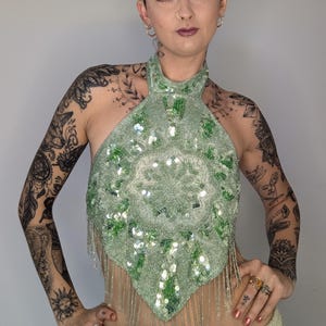 May include: A light green halter top with a beaded design and fringe. The top features a central circular pattern with sequins and beads. The halter top is a unique piece of apparel.