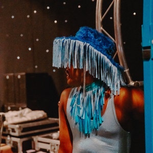 Pu&ograve; includere: Una persona che indossa un cappello blu con frange, una collana a catena d'argento e una canotta blu chiaro.