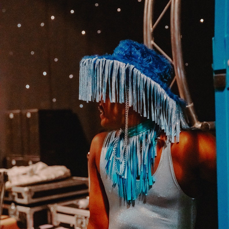 Pu&ograve; includere: Una persona che indossa un cappello blu con frange, una collana a catena d'argento e una canotta blu chiaro.