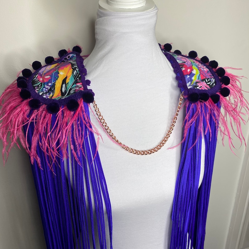 Puede incluir: Boa de plumas morada y rosa con un acento de tela con estampado floral y una cadena dorada. La boa est&aacute; unida a un maniqu&iacute; blanco.