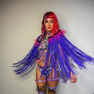 Puede incluir: Una mujer con un top corto de colores y estampado con un chal de flecos morados. El chal tiene plumas rosas en el cuello. Tambi&eacute;n lleva un arn&eacute;s negro con detalles plateados y un par de ligas de muslo con detalles iridiscentes.