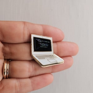Puede incluir: Pin de portátil dorado con pantalla blanca que muestra código. El portátil tiene un teclado blanco y un borde dorado. Un pequeño y detallado accesorio.
