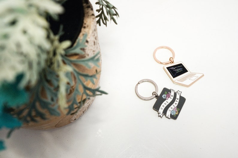 Peut inclure: Deux porte-cl&eacute;s sur le th&egrave;me du codage. Un porte-cl&eacute;s est un ordinateur portable argent&eacute; avec du code noir sur l'&eacute;cran. L'autre porte-cl&eacute;s est un carr&eacute; noir avec une banni&egrave;re blanche qui dit "Code to Create" avec des fleurs sur la banni&egrave;re.