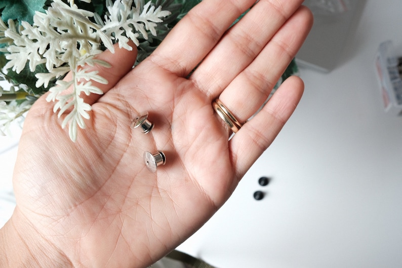 May include: Two silver metal snap fasteners, also known as poppers or press studs, are shown in a hand. The fasteners are commonly used for clothing and other fabric projects.