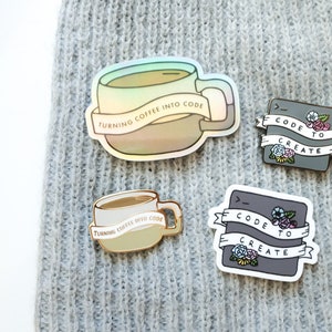 以下が含まれることがあります： コーヒーカップに「Turning coffee into code」という文字が書かれたエナメルピン、グレーの正方形に「Code to create」という文字が書かれたエナメルピン、グレーの正方形に「Code to create」という文字と花が描かれたエナメルピンの3つセットです。