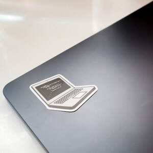 Peut inclure: Un autocollant d'ordinateur portable blanc avec un contour noir, repr&eacute;sentant un ordinateur portable avec du code &agrave; l'&eacute;cran. L'autocollant est plac&eacute; sur un ordinateur portable gris fonc&eacute;.