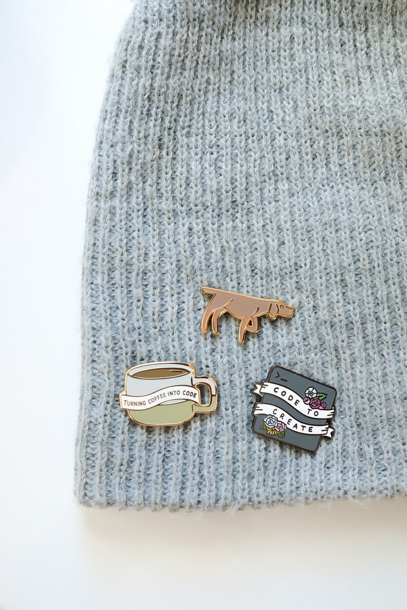 May include: Three enamel pins on a grey knitted beanie. The pins are gold-toned and feature a coffee cup with the text "Turning coffee into code", a dog, and a black rectangle with the text "Code to create" and floral accents.