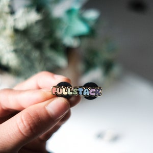 May include: A rainbow enamel pin with the word "FULLSTACK" in gold lettering. The pin is attached to a black backing.