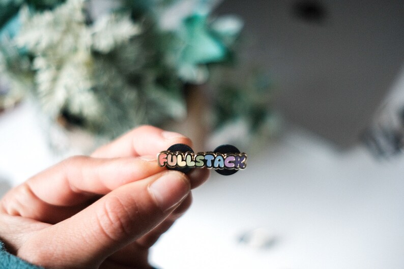 May include: A rainbow enamel pin with the word "FULLSTACK" in gold lettering. The pin is attached to a black backing.