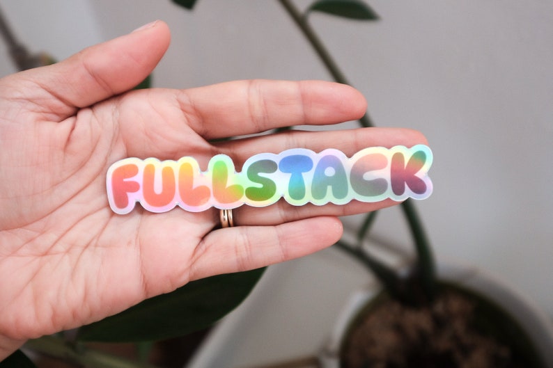 K&ouml;nnte beinhalten: Ein holografischer Aufkleber mit dem Wort "FULLSTACK" in Regenbogenfarben. Der Aufkleber befindet sich auf der Hand einer Person.