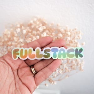 K&ouml;nnte beinhalten: Ein holografischer Aufkleber mit dem Text "FULLSTACK" in einem Regenbogen-Farbschema. Der Aufkleber wird in einer Hand gehalten.