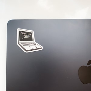 Peut inclure: Un ordinateur portable noir avec un autocollant repr&eacute;sentant un ordinateur portable affichant du code sur l'&eacute;cran. L'ordinateur portable a un logo Apple argent&eacute; au dos.