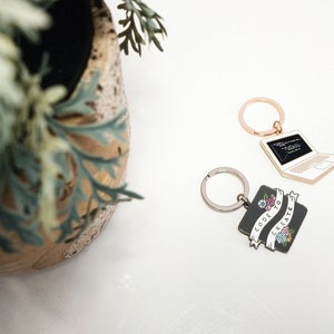 Pu&ograve; includere: Due portachiavi a tema coding. Un portachiavi &egrave; un laptop argentato con codice nero sullo schermo. L'altro portachiavi &egrave; un quadrato nero con uno striscione bianco che dice "Code to Create" con fiori sullo striscione.