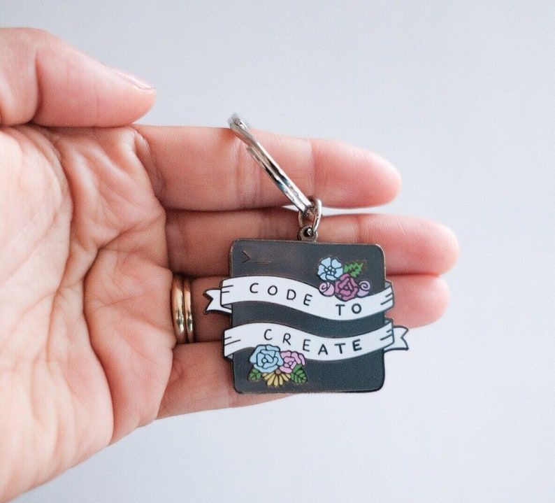 Pu&ograve; includere: Un portachiavi in metallo argentato con un ciondolo quadrato nero. Il ciondolo ha un testo bianco che dice "CODE TO CREATE" con un design floreale.
