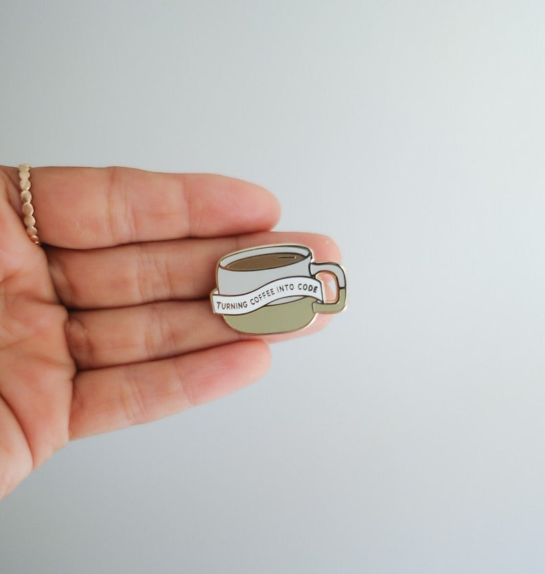 以下が含まれることがあります： 白いバナーに「Turning coffee into code」という文字が書かれたコーヒーカップが描かれた金色のエナメルピンです。