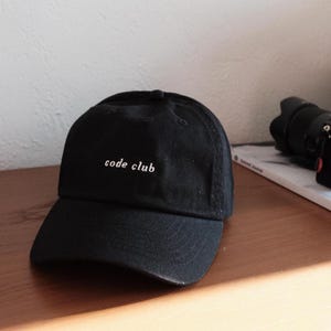 May include: A black baseball cap with the words "code club" embroidered in white. The cap features a curved peak and is made from a soft material. The hat is on a wooden surface.