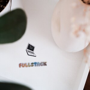 K&ouml;nnte beinhalten: Eine wei&szlig;e Oberfl&auml;che mit einem schwarz-wei&szlig;en Aufkleber eines Laptops mit Code auf dem Bildschirm. Unter dem Laptop befindet sich ein Aufkleber mit der Aufschrift "FULLSTACK" in Regenbogenfarben.