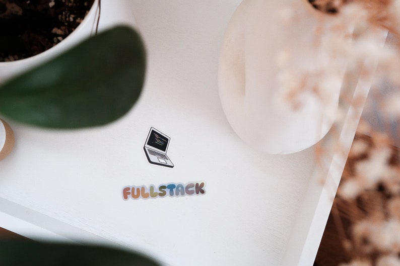 K&ouml;nnte beinhalten: Eine wei&szlig;e Oberfl&auml;che mit einem schwarz-wei&szlig;en Aufkleber eines Laptops mit Code auf dem Bildschirm. Unter dem Laptop befindet sich ein Aufkleber mit der Aufschrift "FULLSTACK" in Regenbogenfarben.