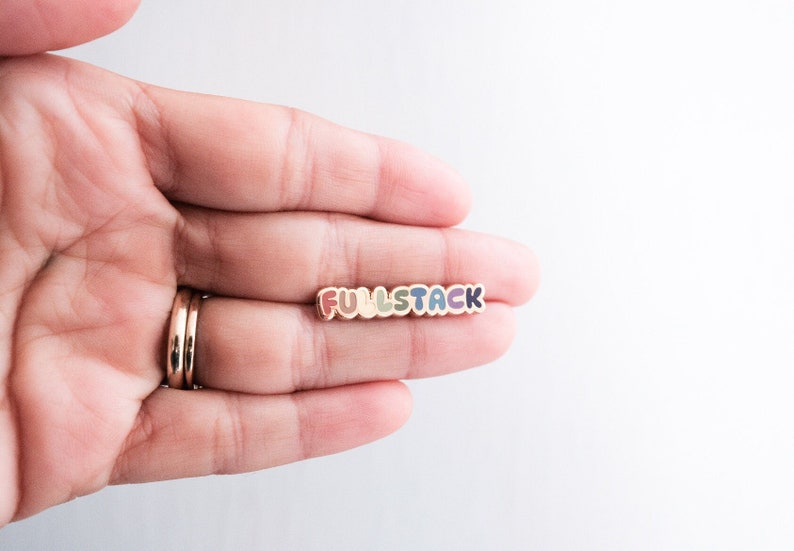 May include: A colorful enamel pin with the word "FULLSTACK" in rainbow colors. The pin is held in a hand.