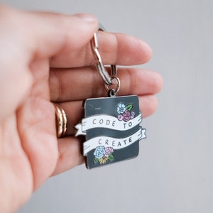 Pu&ograve; includere: Un portachiavi in metallo nero con uno striscione bianco che recita "CODE TO CREATE" e un design floreale.