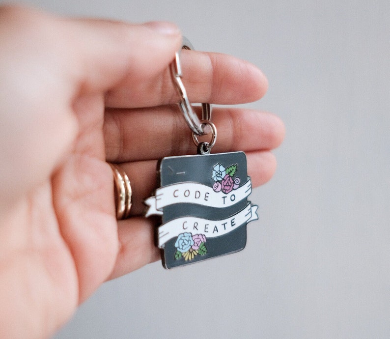 Pu&ograve; includere: Un portachiavi in metallo nero con uno striscione bianco che recita "CODE TO CREATE" e un design floreale.
