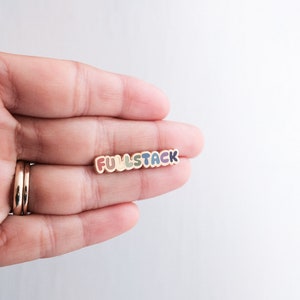 May include: A colorful enamel pin with the word "FULLSTACK" in rainbow colors. The pin is held in a hand.