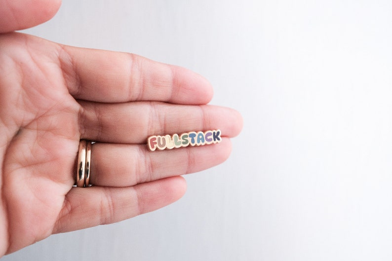 May include: A colorful enamel pin with the word "FULLSTACK" in rainbow colors. The pin is held in a hand.