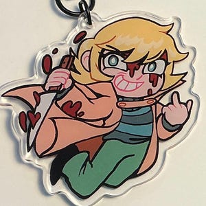 以下が含まれることがあります： 透明なアクリルキーホルダー。金髪のキャラクターが描かれており、ベージュのコート、青と緑のストライプのシャツ、緑のズボンを着用しています。キャラクターは赤い血の飛沫がついたナイフを持ち、手でジェスチャーをしています。