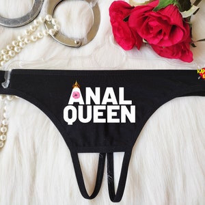 May include: Black thong underwear with the text "ANAL QUEEN" in white block letters. The design includes a crown and a pink flower graphic. The underwear is displayed on a hanger.