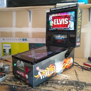 May include: A virtual pinball machine with a black and gray cabinet. The machine features a screen displaying "ELVIS" in large red letters. The cabinet has "Bally Pinball" branding and colorful graphics.