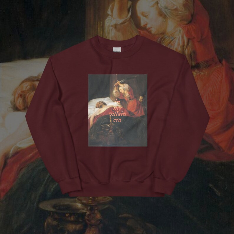 May include: Burgundy sweatshirt with a painting of a woman stabbing a man in bed. The text "in my villain era" is printed in pink on the painting.