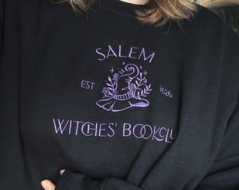 Puede incluir: Una sudadera negra de cuello redondo con bordado morado en el pecho. El diseño incluye un sombrero de bruja rodeado de elementos florales, estrellas y el texto SALEM EST 1626, con las palabras WITCHES' BOOKCLUB bordadas bajo el gráfico.