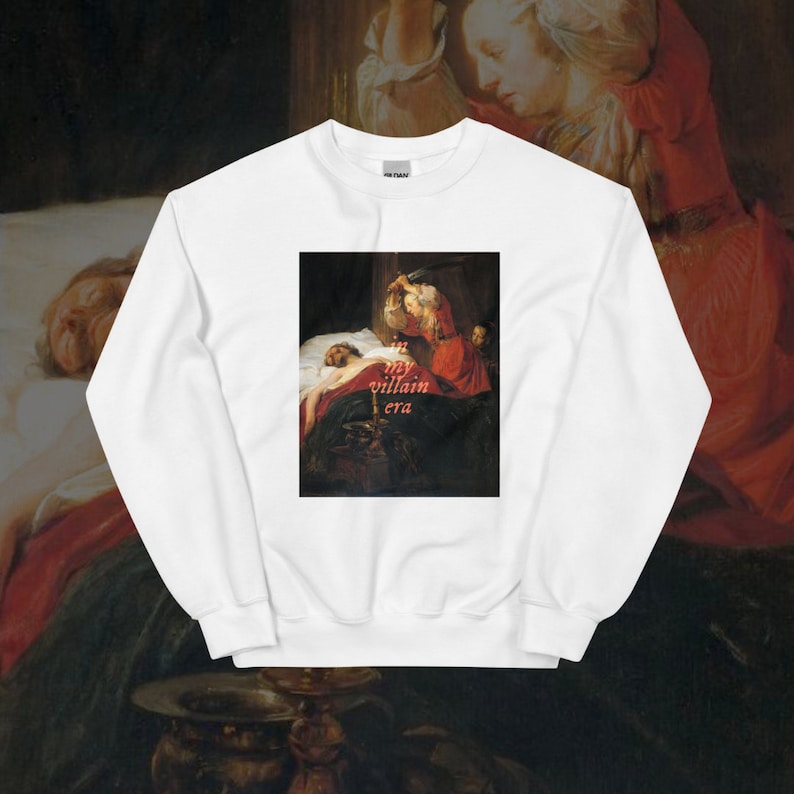 May include: White sweatshirt with a painting of a woman holding a knife over a man in bed. The text "my villain era" is printed in pink on the painting.