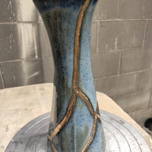 Puede incluir: Un jarrón de cerámica azul y blanco con reparaciones doradas estilo kintsugi. El jarrón tiene una parte superior acampanada y una base redondeada. Las líneas doradas crean un patrón decorativo.