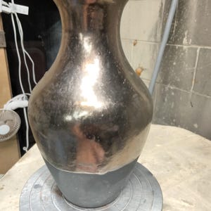 May include: A ceramic vase with a flared rim and a tapered neck. The upper portion of the vase features a reflective, metallic silver finish, while the lower portion has a matte, dark gray finish with a visible horizontal transition line.
