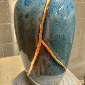 May include: A ceramic vase with a glossy blue and dark-toned glaze, featuring prominent gold-colored repair lines that trace cracks across the surface. The vessel has a rounded body and a narrow opening at the top, showcasing a smooth finish.