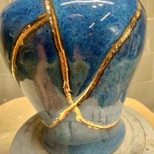 May include: A ceramic vase with a glossy blue glaze featuring prominent gold-colored veins that crisscross the surface. The vase has a rounded body that tapers toward the base and a short, narrow neck with a gold-colored rim at the top opening.