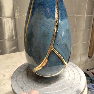 May include: A teardrop-shaped ceramic vase with a glossy, mottled blue glaze. The surface features prominent, raised gold-colored lines that create a branching, crack-like pattern across the body of the vessel, extending from the top to the base.