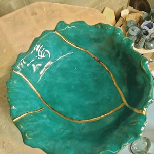 May include: A handcrafted, teal-colored ceramic bowl with a leaf-like shape. The bowl features a gold-colored line design and a glossy finish. The bowl's edges are wavy, adding to its organic aesthetic. This decorative bowl is suitable for serving or display.