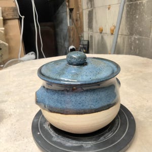 Peut inclure: Un pot en céramique fait à la main avec un couvercle. Le pot est de couleur beige clair en bas, avec une bande bleu foncé et un couvercle bleu moucheté. Le pot a deux petites poignées et un petit bouton sur le couvercle.