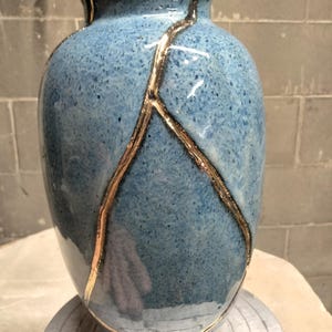 May include: A blue ceramic vase with a speckled finish and a narrow neck. The surface features prominent, raised gold-colored lines that branch out from the top rim down toward the base, creating a distinct decorative pattern across the body.