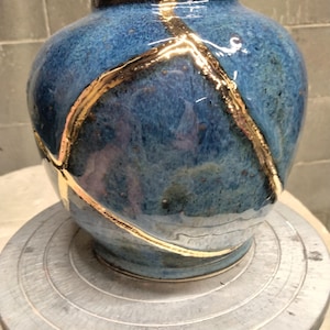May include: A rounded ceramic vase with a glossy blue glaze featuring a network of cracks repaired with a metallic gold finish. The vessel has a narrow neck and a wide body, showcasing a distinct contrast between the deep blue and the gold lines.
