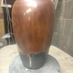 May include: A tall, rounded ceramic vase featuring a two-tone glaze. The upper portion is a warm, mottled brown, while the lower section is a solid matte black. The glaze transition creates a distinct drip effect where the brown overlaps the black.