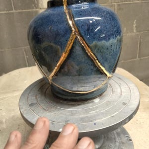 May include: A small, rounded ceramic vase with a glossy, deep blue glaze. The surface features prominent, jagged cracks repaired with a metallic gold lacquer, highlighting the broken sections in a distinct, contrasting pattern across the body.