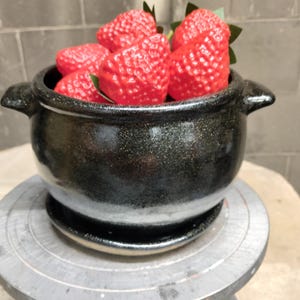 May include: A black ceramic pot with a speckled finish and two side handles, filled with several bright red artificial strawberries. The pot sits on a matching black ceramic saucer, creating a contrast between the dark vessel and the fruit.