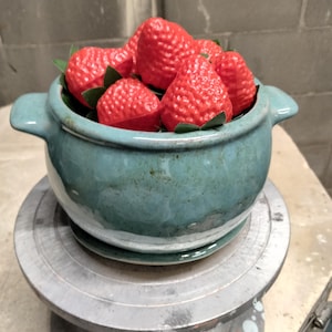 May include: A round, teal ceramic bowl with two side handles, filled with several bright red, textured strawberry-shaped objects. The bowl features a glossy, mottled glaze and sits on a matching circular base, containing green leaf accents.