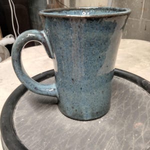 May include: A blue ceramic mug with a speckled glaze and a rounded handle. The mug is sitting on a black surface.