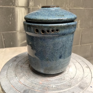 Può includere: Un barattolo cilindrico in ceramica con smalto blu maculato e coperchio abbinato. La parte superiore del barattolo presenta una fila orizzontale di cinque piccoli fori di ventilazione circolari. La superficie ha una finitura lucida.