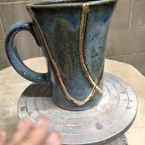 May include: A ceramic mug with a mottled blue and grey glaze featuring a prominent gold-colored crack repair design that runs vertically along the side. The mug has a curved handle and a wide rim, showcasing a smooth, glossy finish throughout.