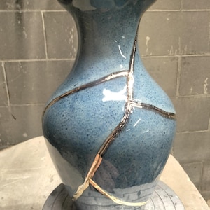 May include: A blue ceramic vase with a glossy, speckled finish featuring prominent gold-colored repair lines running across the body. The vase has a wide, flared rim and a tapered neck, resting on a circular base with a smooth, reflective surface.